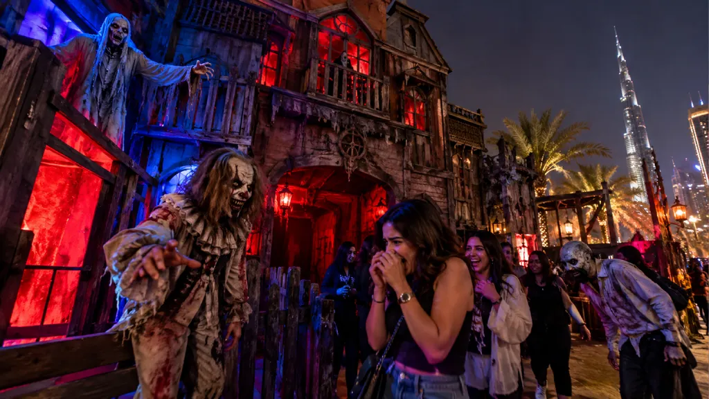 Haunted house attraction in Dubai at night, featuring costumed horror actors, a gothic mansion illuminated with red and blue lights, and frightened visitors reacting to Halloween scares, with the Burj Khalifa visible in